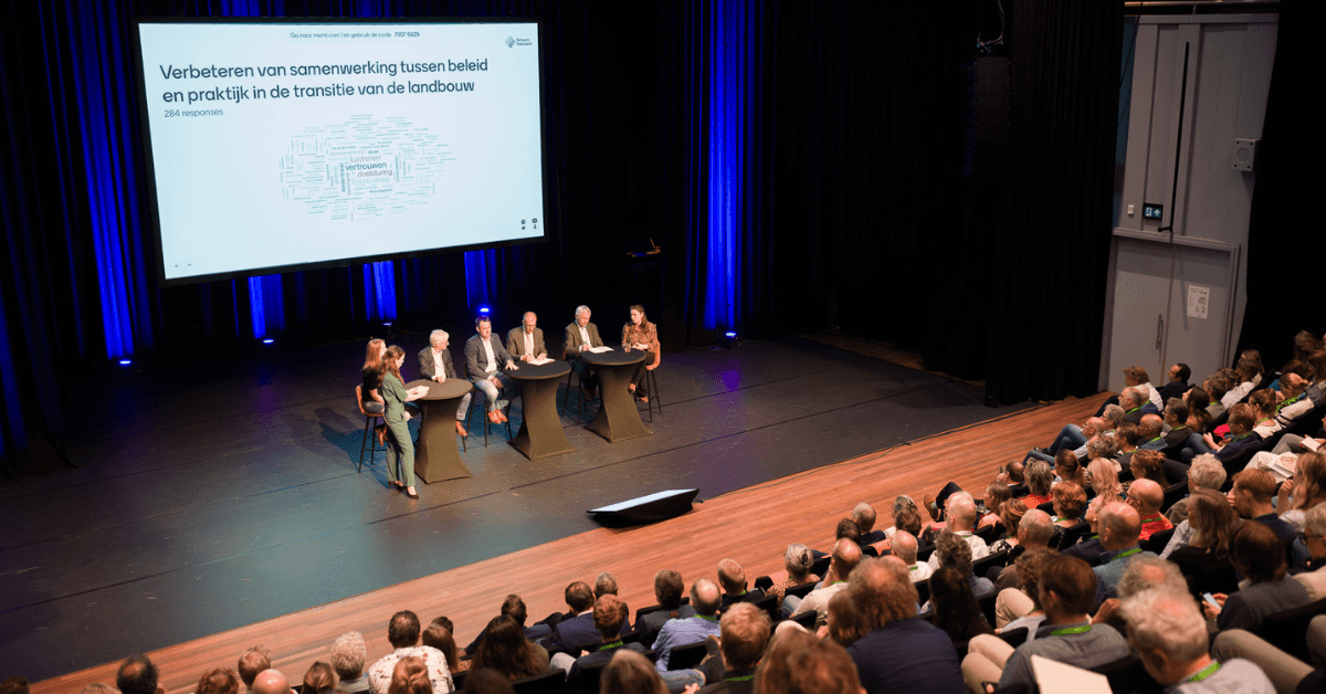 Overzichtsbeeld van een volle theaterzaal met panelleden op het podium