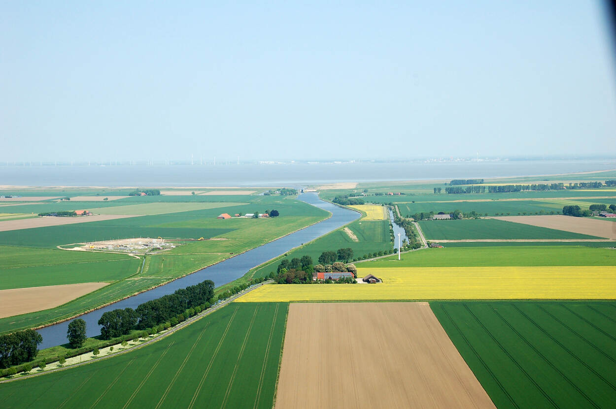 Foto van landschap Binnen AA richting Nieuw Statenzijl, Noord-Oost Groningen met akkers op de voorgrond, vanaf linksvoor loopt een rivier dat in de verte uitmondt in de waddenzee met in de verte aan de horizon windmolens. In het midden van de pagina staat een oude Groningse boerderij omringd door bomen.